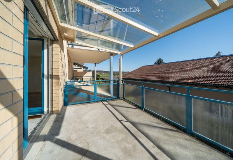 Balcony / Terrace. Spacious balcony with a modern glass roof and blue railings, overlooking a residential area with a clear blue sky.