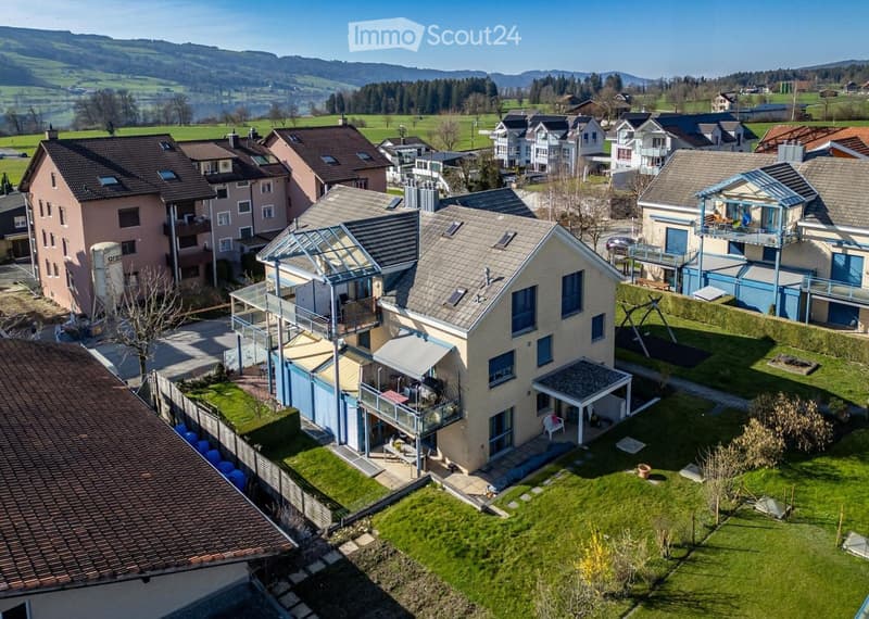 House. Aerial view of a residential area with multiple houses, surrounded by green fields and distant hills.
