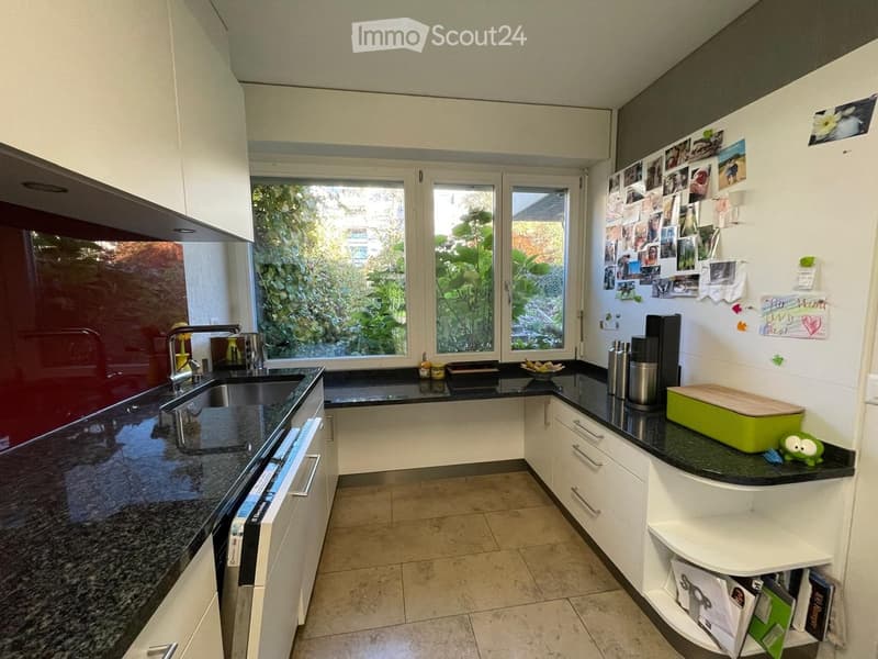 Modern kitchen with white cabinets, black granite countertops, stainless steel sink, and various appliances. Features a window with a view of greenery, a built-in oven, and a small shelf with magazines.