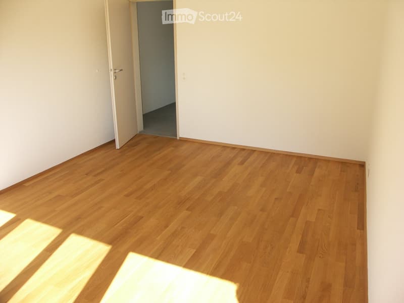 Empty room with light wooden flooring and white walls, featuring an open doorway leading to another space.