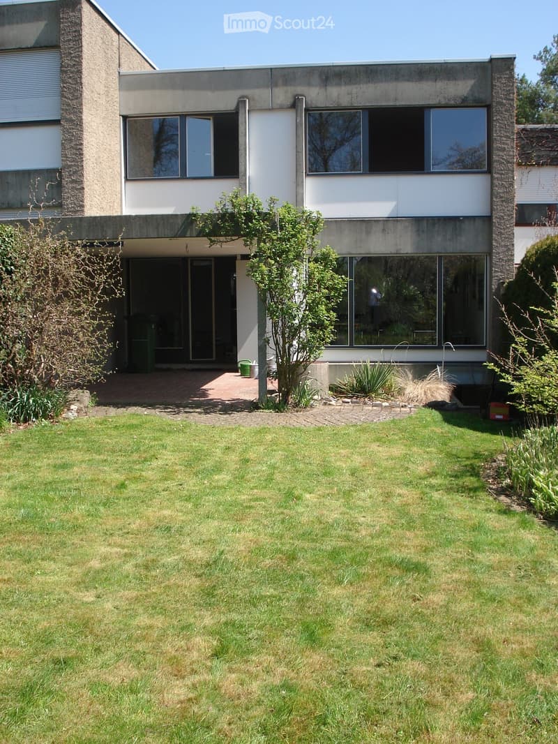 Modern two-story house with large windows, concrete facade, and a well-maintained lawn in front.