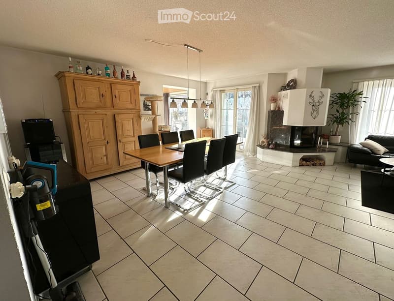 Open-plan living and dining area with tiled flooring, a large wooden dining table with chairs, a black leather sofa, a wooden cabinet, a fireplace with a decorative deer head, and a potted plant near the window.