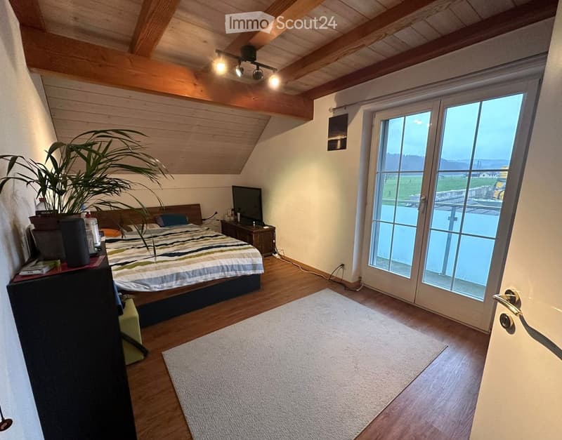 Cozy bedroom with wooden floors, slanted wooden ceiling, large window with view, bed with striped bedding, nightstand, TV, and plant.