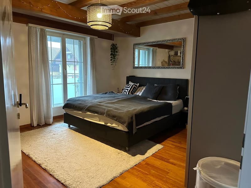 Cozy bedroom with wooden floors, a large bed with dark bedding, a nightstand, a large mirror, and a pendant light fixture.