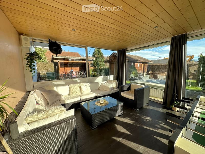 A modern, enclosed patio with a wooden ceiling, large windows, and a comfortable seating area featuring a sofa, coffee table, and chairs. The space includes a small bar area with a foosball table and is decorated with plants and a hanging basket.