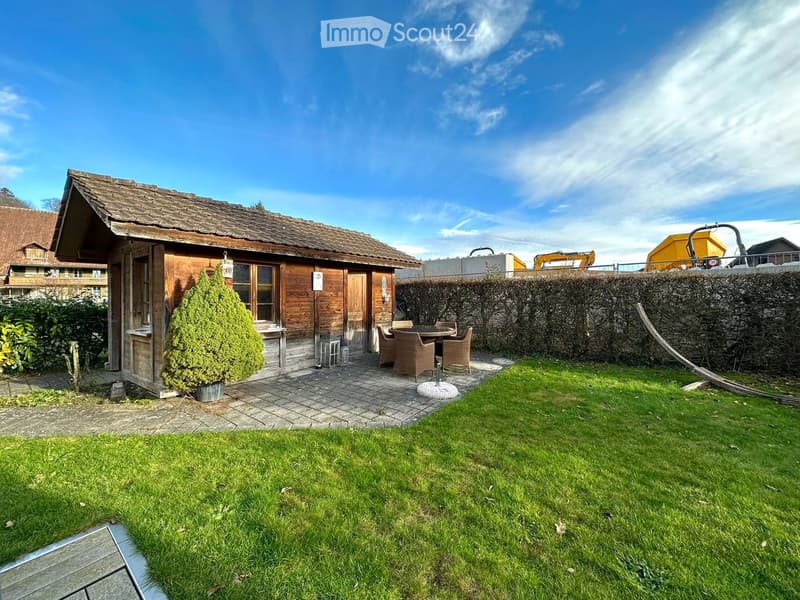A wooden house with a tiled roof, surrounded by a well-maintained garden with a patio area featuring a table and chairs, a small tree, and a stone wall.
