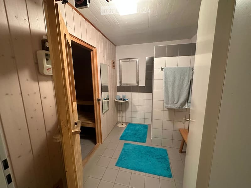 Bathroom with tiled walls and floor, a small sink area with a mirror, a towel hanging on a rack, and a wooden bench near the shower area.