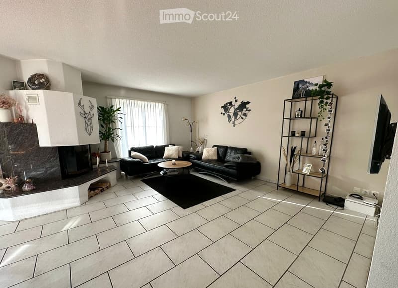 Modern living room with a black leather sofa, coffee table, fireplace, large window with white curtains, and a shelf with decorative items.
