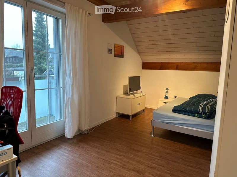 Cozy bedroom with wooden floors, slanted wooden ceiling, large window with white curtains, bed with striped blanket, nightstand with lamp, small cabinet with screen, and red mesh chair.