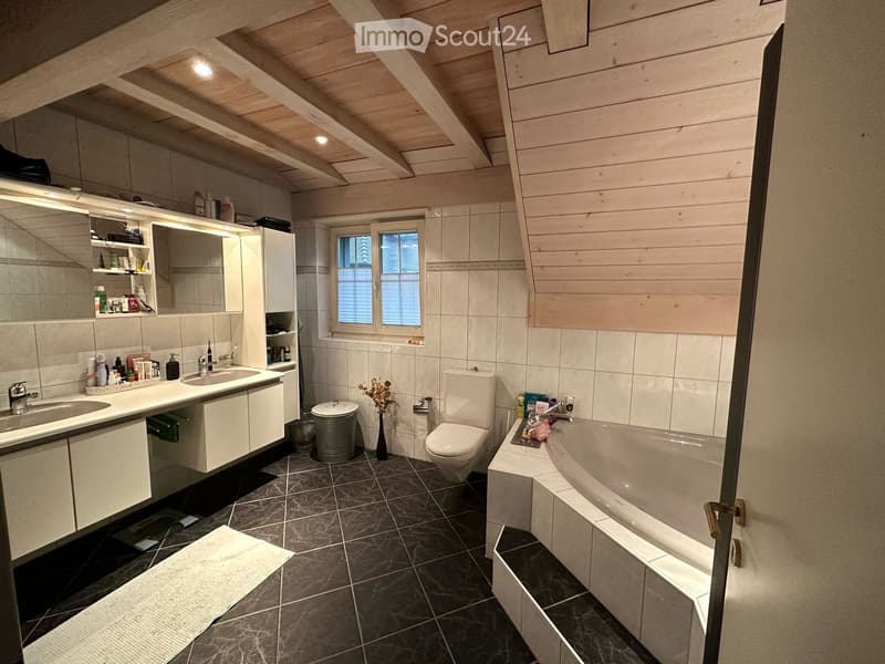 Modern bathroom with dark tiled floor, white cabinets, double sink, toilet, bathtub, and a large mirror. Features wooden slanted ceiling and a window with blinds.