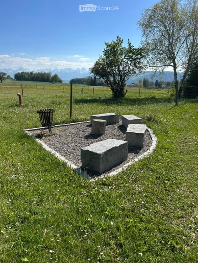 Garden / Outdoor Area. Outdoor seating area with concrete benches arranged in a semi-circle on a gravel surface, surrounded by grass and trees, with a scenic mountain view in the background.