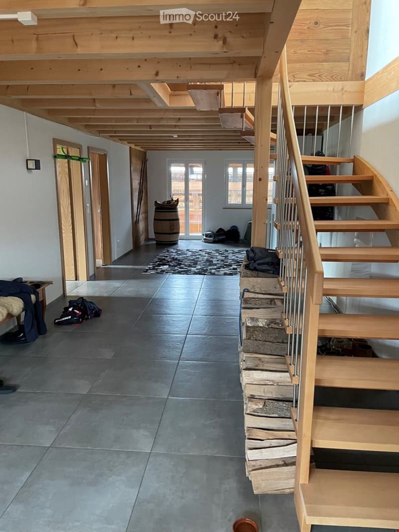 Corridor / Hallway. Spacious hallway with exposed wooden beams, tiled floor, staircase leading upwards, and scattered items including bags and a barrel.