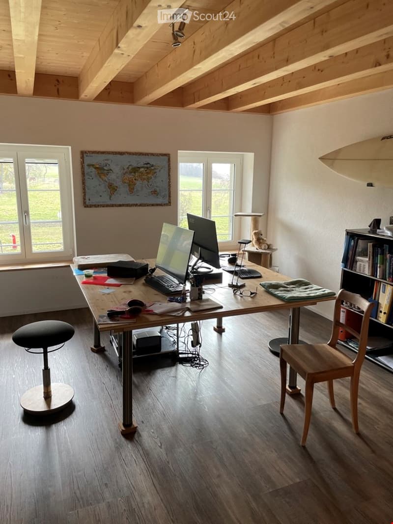 Furnished Room. Bright, spacious room with wooden flooring and exposed wooden beams on the ceiling. Features a large desk with a laptop, monitor, keyboard, mouse, and various office supplies. A wooden chair and a black swivel stool are present. A cat tree