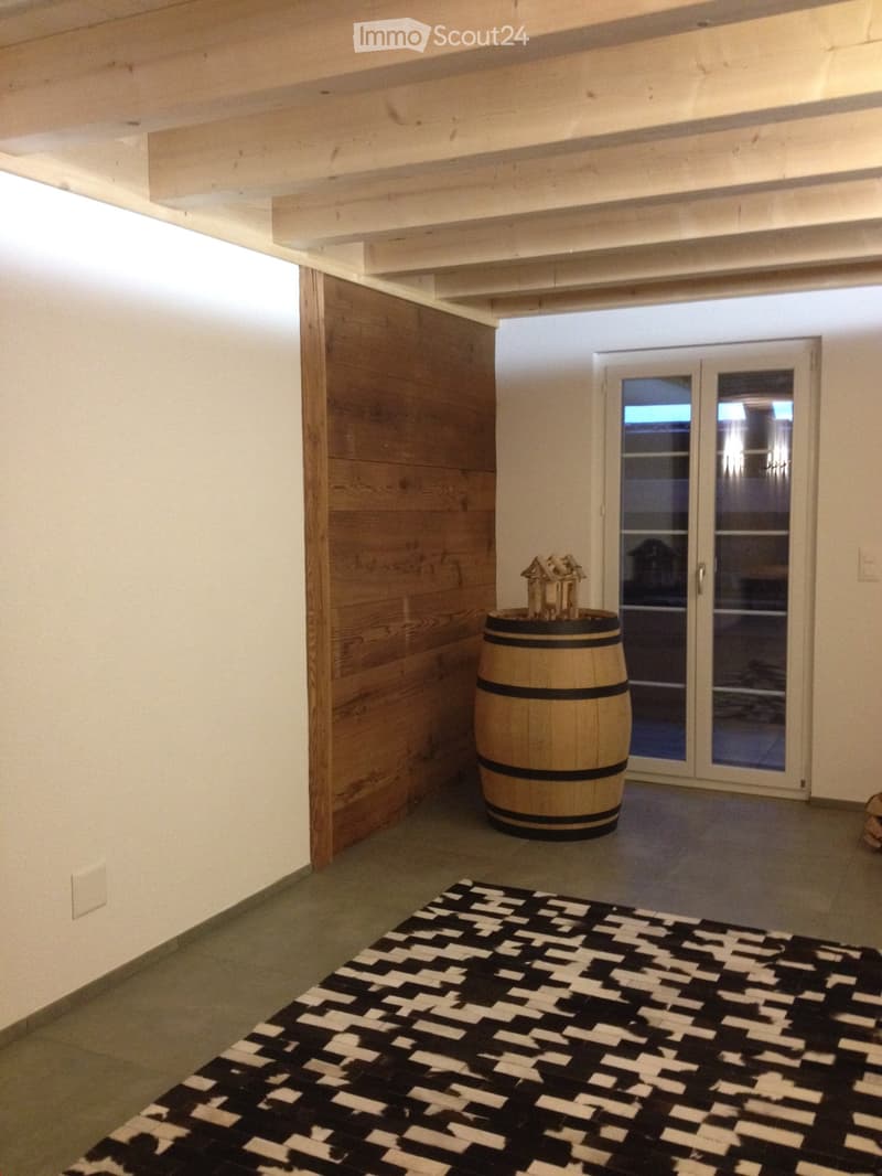Furnished Room. A small, well-lit room with exposed wooden beams, a patterned black and white rug, a wooden wine barrel with a miniature house on top, and a glass door leading outside.