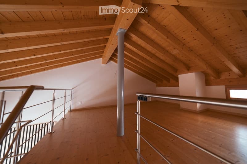 An empty attic space with wooden beams and a wooden floor, featuring a metal railing and a central support column.