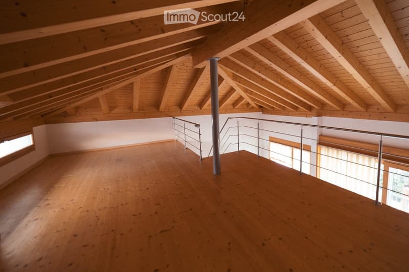 Empty room with wooden floor and exposed wooden beams on the ceiling, featuring a metal railing and a central support column.