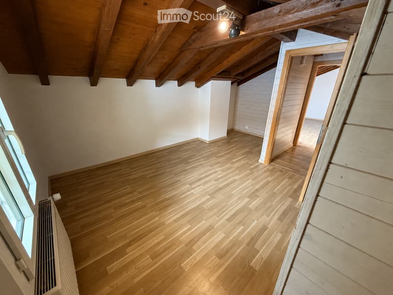 Empty room with wooden flooring, white walls, exposed wooden beams on the ceiling, and a large window on the left side.