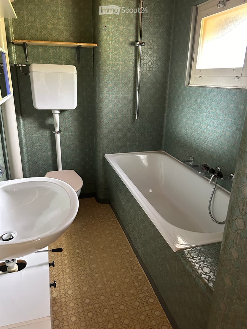 Bathroom with green patterned walls, a white toilet, a bathtub with a handheld shower, and a sink. The floor has a patterned tile design.