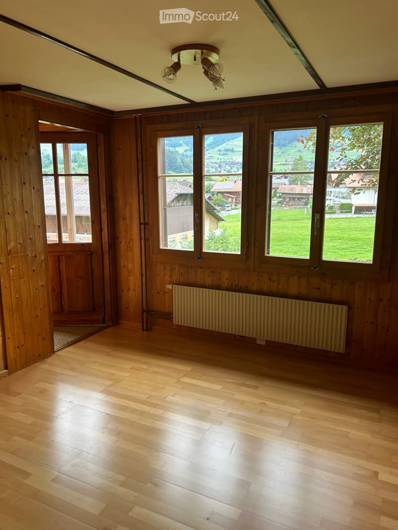 Empty room with wooden walls and floor, large windows offering scenic views, and a radiator below one window.