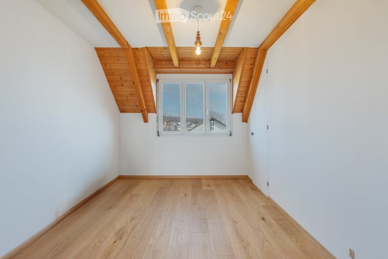 Empty room with wooden flooring, white walls, and exposed wooden beams on the ceiling. A large window with a view of the outside is present.