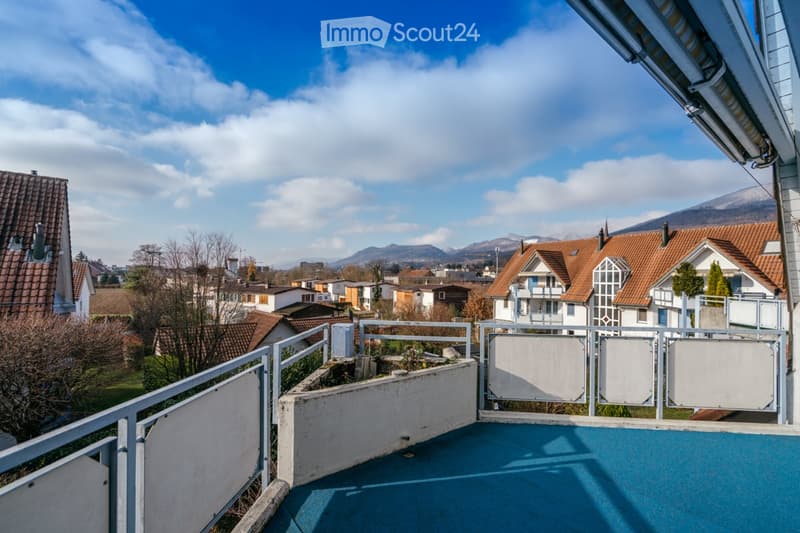 Balcony with metal railing, overlooking a residential area with houses and distant mountains under a partly cloudy sky.