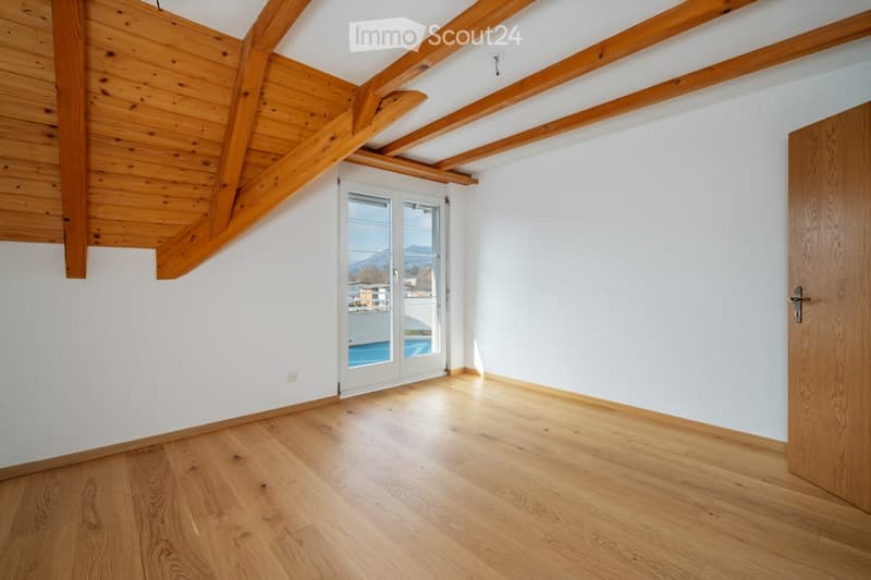 Empty room with wooden flooring, white walls, exposed wooden beams on the ceiling, and a large window offering a scenic view.