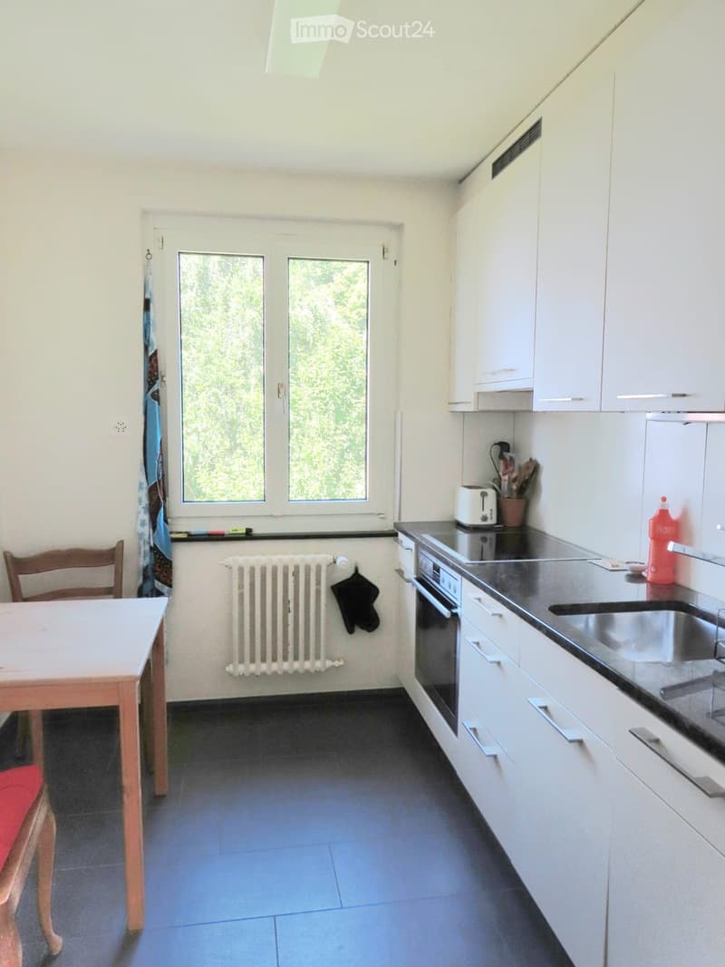 Modern kitchen with white cabinets, dark countertops, and a small dining area. Features include a sink, oven, toaster, and a small table with a chair.