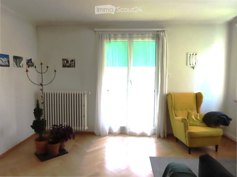 Bright living room with wooden flooring, large window with white curtains, yellow armchair, potted plants, candelabra, radiator, and picture frames on the wall.