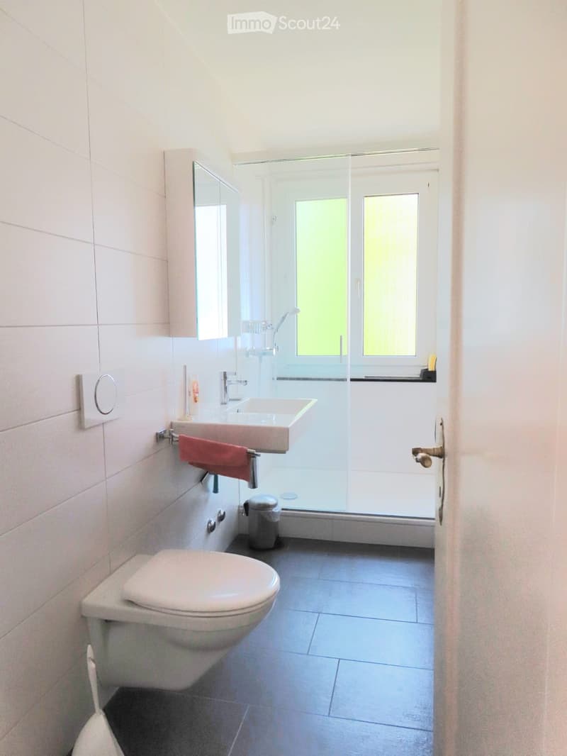 Modern bathroom with white tiles, a toilet, a sink with a mirror, a towel, and a glass-enclosed shower area.
