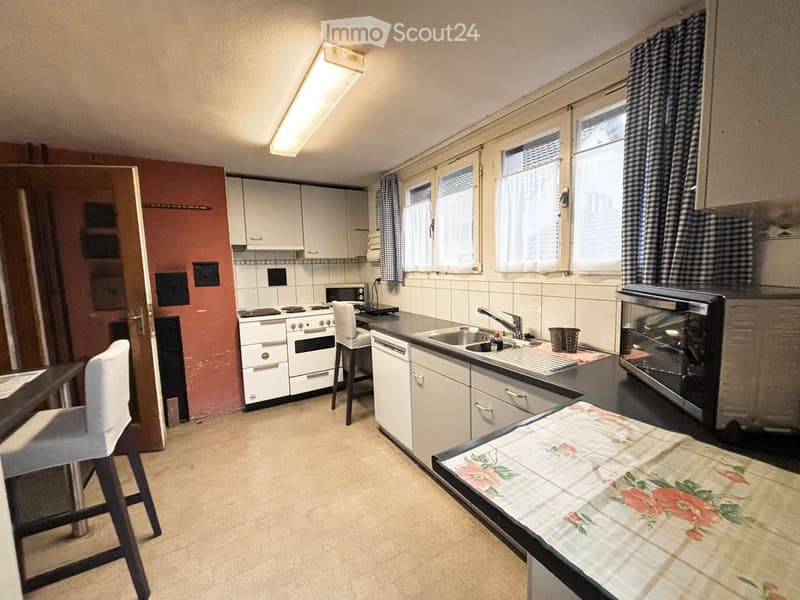 A compact kitchen with white cabinets, a stove, sink, microwave, and a small table with a floral tablecloth. The space includes a window with checkered curtains and a chair near the stove.