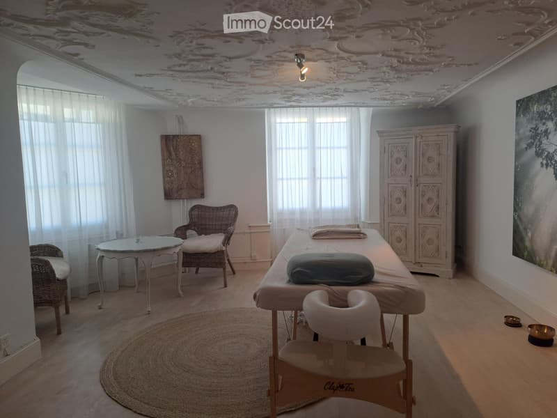 Well-lit room with a massage table, two wicker chairs, a small round table, a cabinet, and large windows with white curtains.