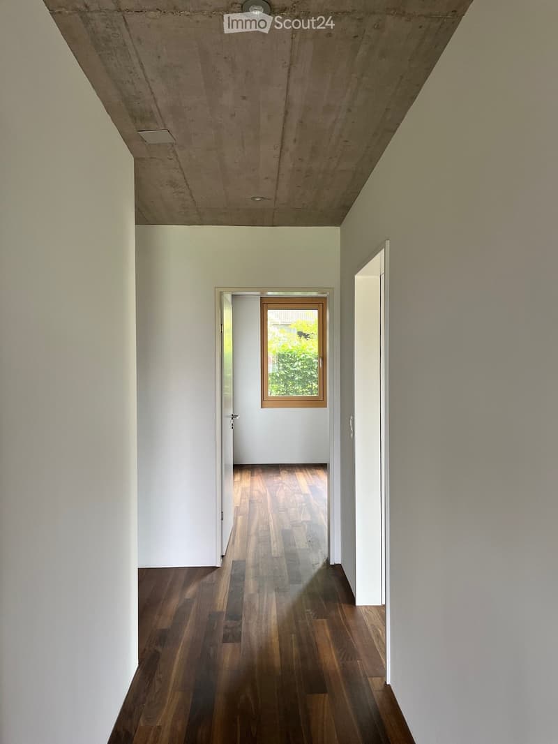 Empty hallway with wooden flooring, white walls, and a door leading to another room with a window showing greenery outside.