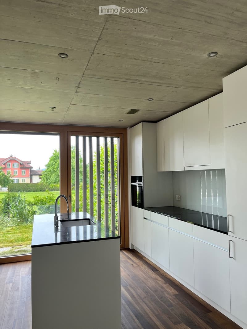 Modern kitchen with white cabinets, dark countertops, and a large island with a sink and faucet. The space features wooden flooring and large windows offering a view of the garden.