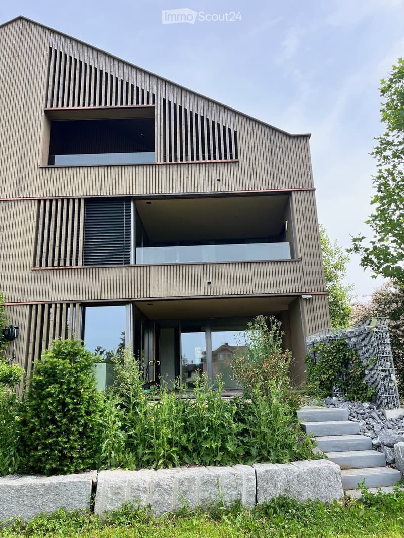 Modern two-story house with wooden exterior, large windows, and balconies. Features include a raised ground floor, steps leading up to the entrance, and surrounding greenery.