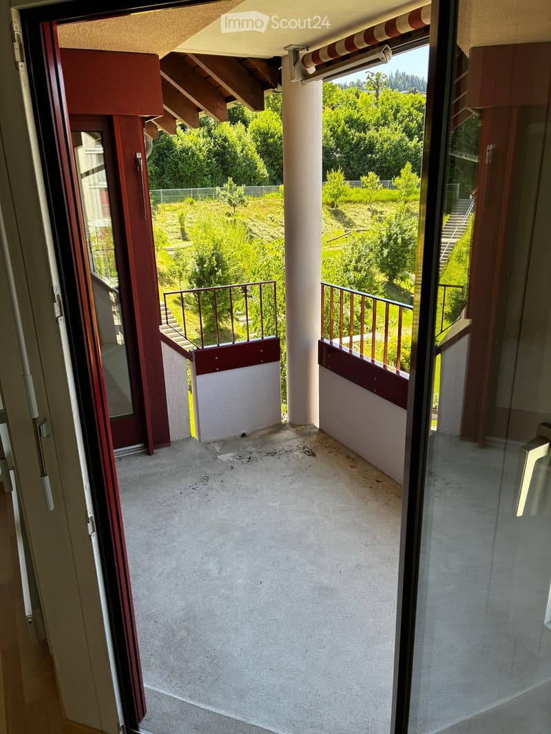 Balcony with concrete floor, metal railings, and wooden handrails, overlooking a green landscape with trees and hills.