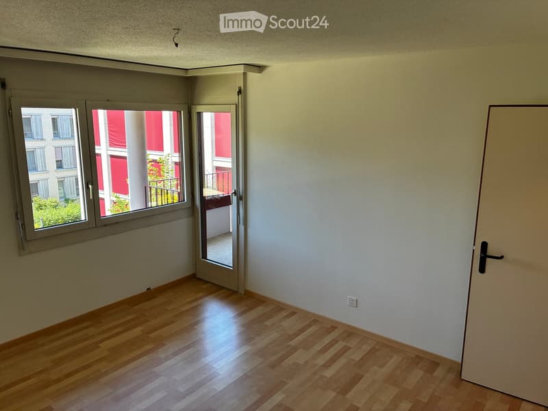 Empty room with wooden flooring, white walls, and a large window with a sliding door leading to a balcony area.