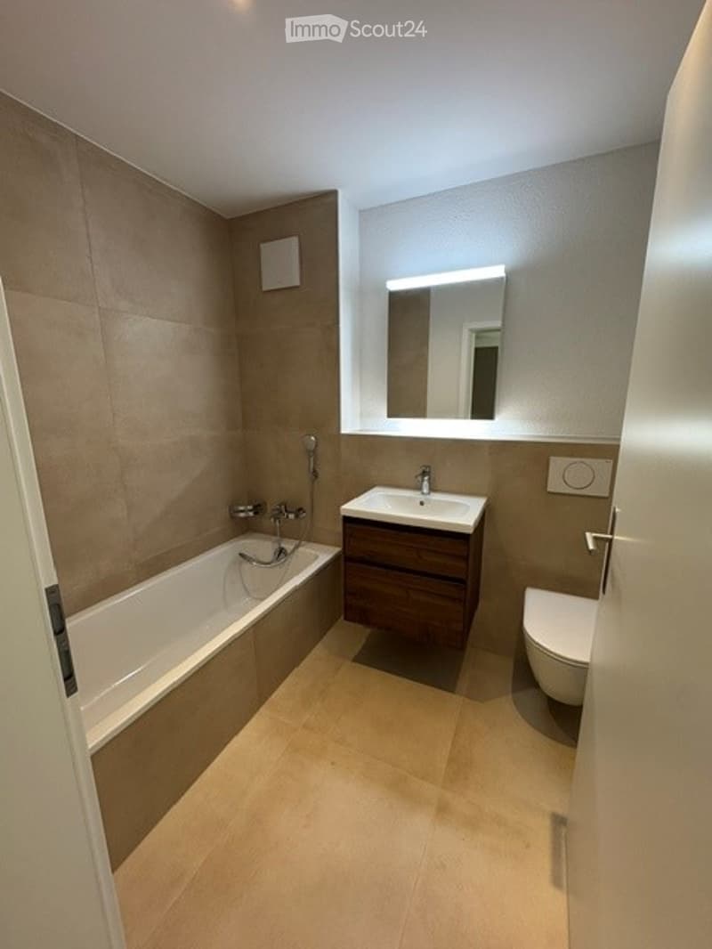 Modern bathroom with a bathtub, sink, toilet, and a large mirror above the sink. The walls are tiled in a neutral color, and the floor is covered with light-colored tiles.