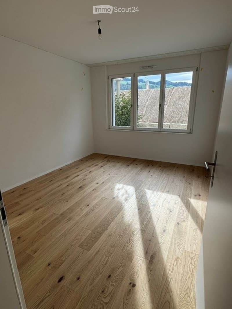 Empty room with wooden flooring and large window allowing natural light to enter.