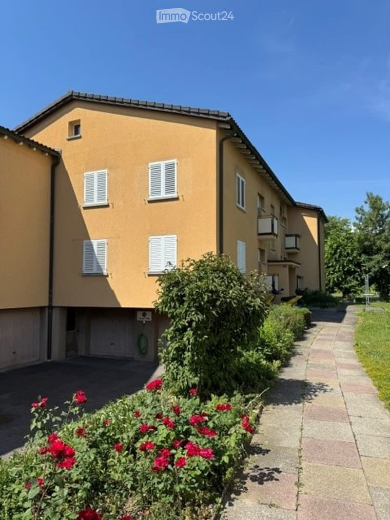 Two-story residential building with beige facade, white shutters, and a garage entrance. Bushes and red flowers line the pathway.