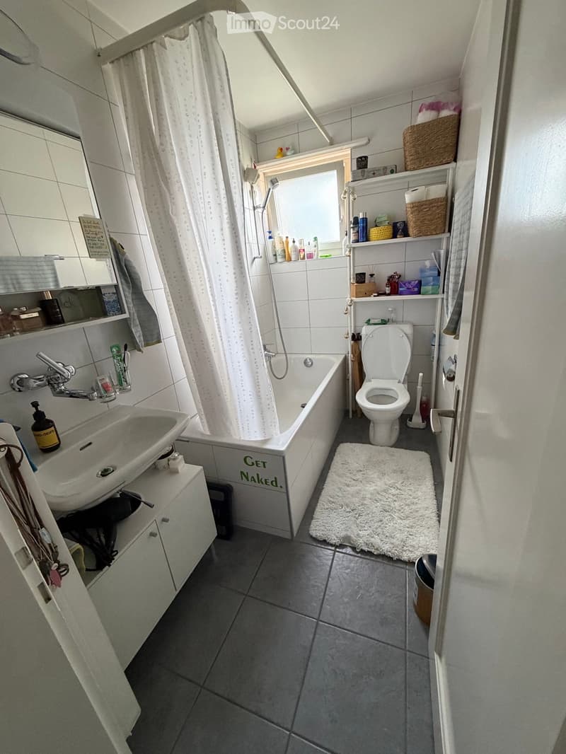 Bathroom with a white tiled shower area, bathtub, toilet, sink, shelves, and a white rug on the floor.