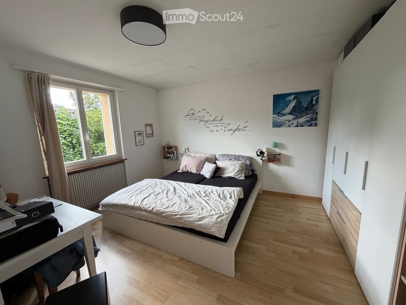 Cozy bedroom with a double bed, white bedding, a small table with a laptop, a chair, and a large window with curtains. The room features wooden flooring, a wardrobe, and decorative wall art.