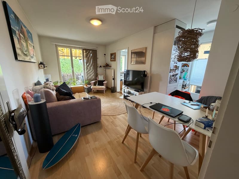 A modern living room with a sofa, a dining table with chairs, a laptop, a surfboard, a television, and a floor lamp. The room features wooden flooring and large windows with a view of greenery.