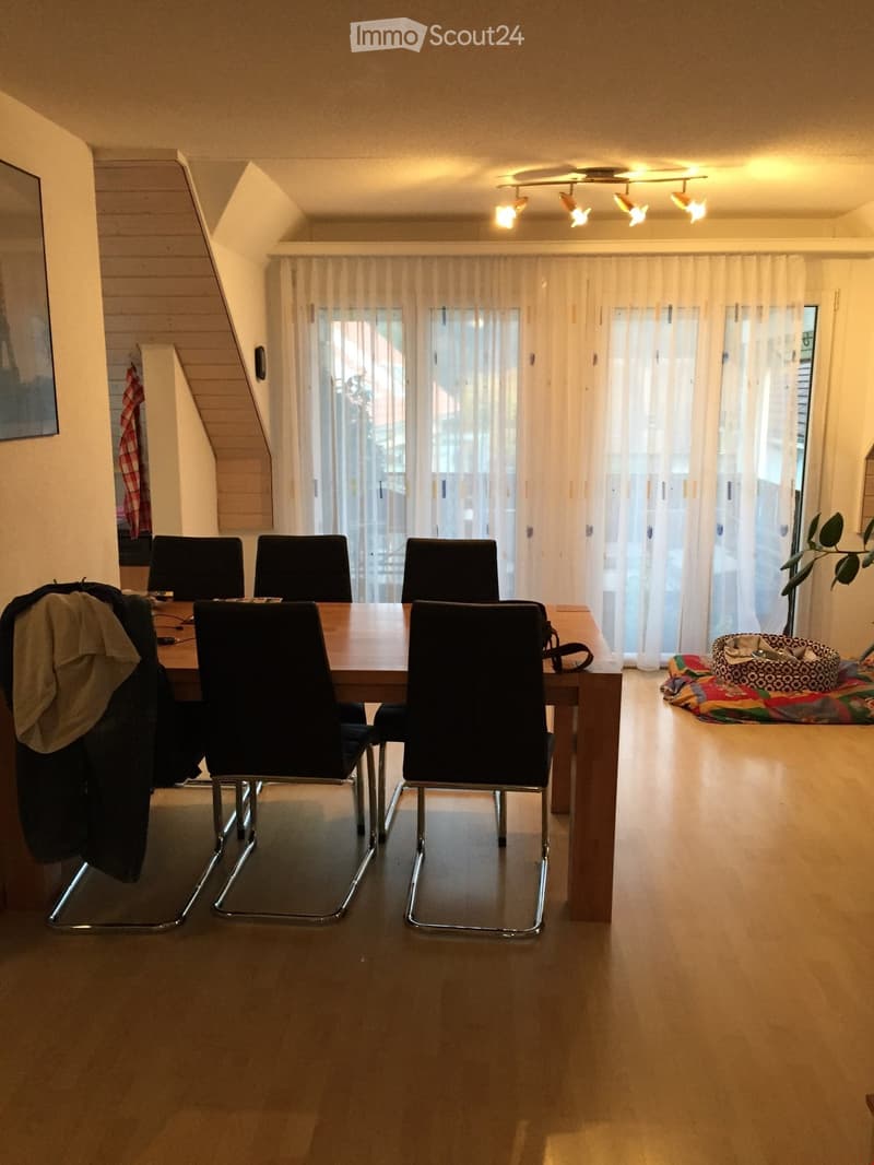 Dining room with a wooden table, four black chairs, large windows with white curtains, and a small plant in a basket on the floor.