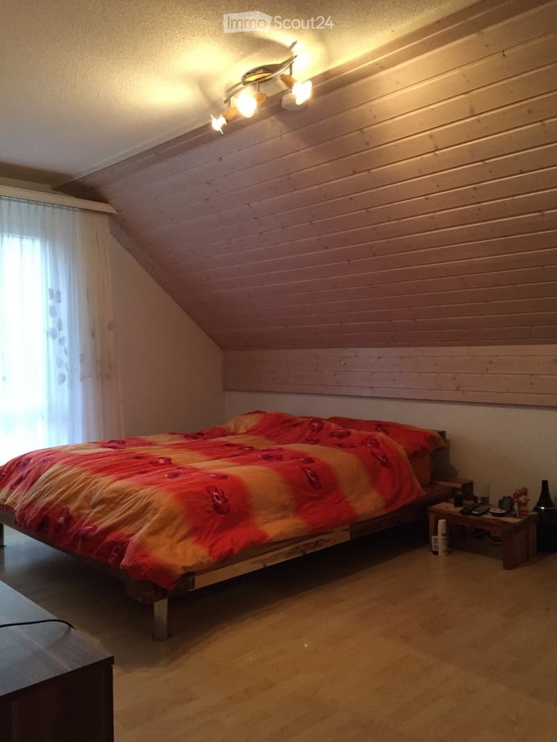 Cozy attic bedroom with wooden slanted ceilings, a bed with a red and orange floral duvet, a nightstand with a remote control and bottle, and a window with white curtains.