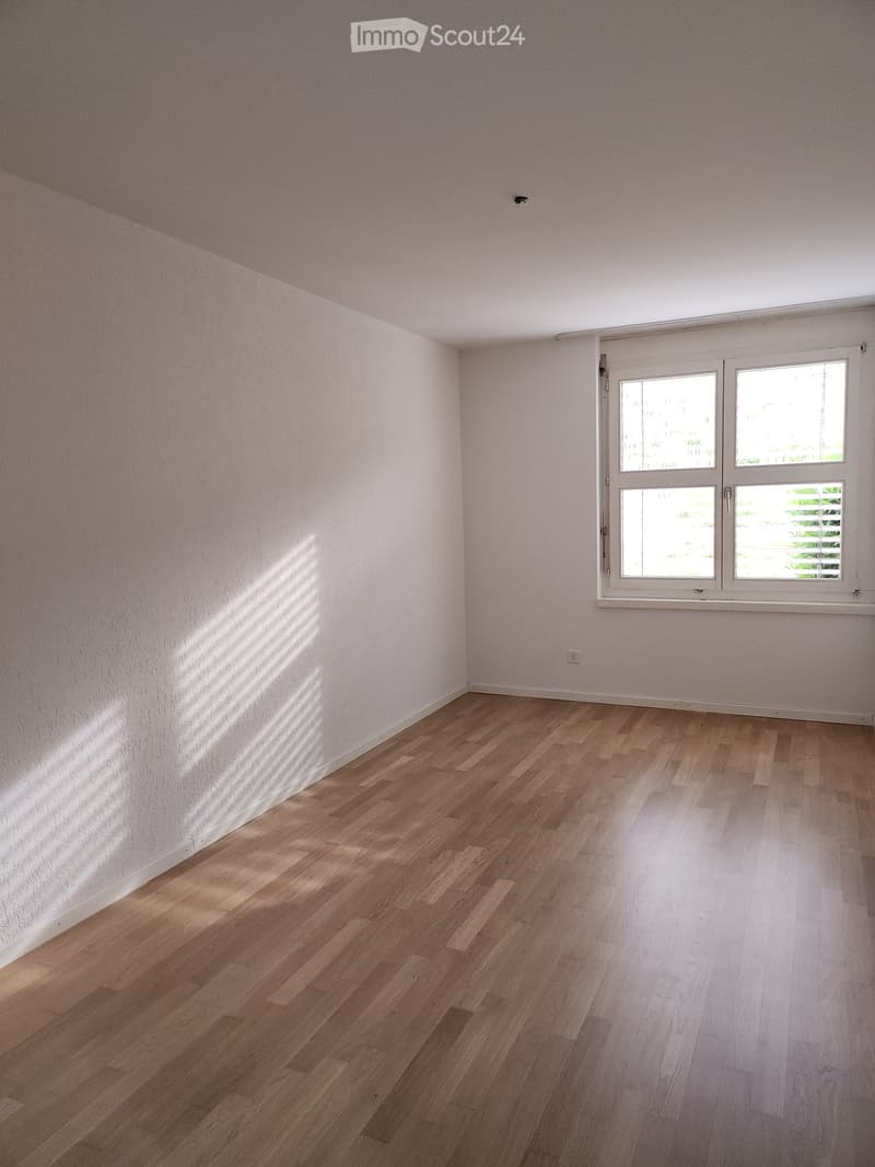 Empty room with light wooden flooring and white walls, featuring a large window with blinds allowing natural light to enter.