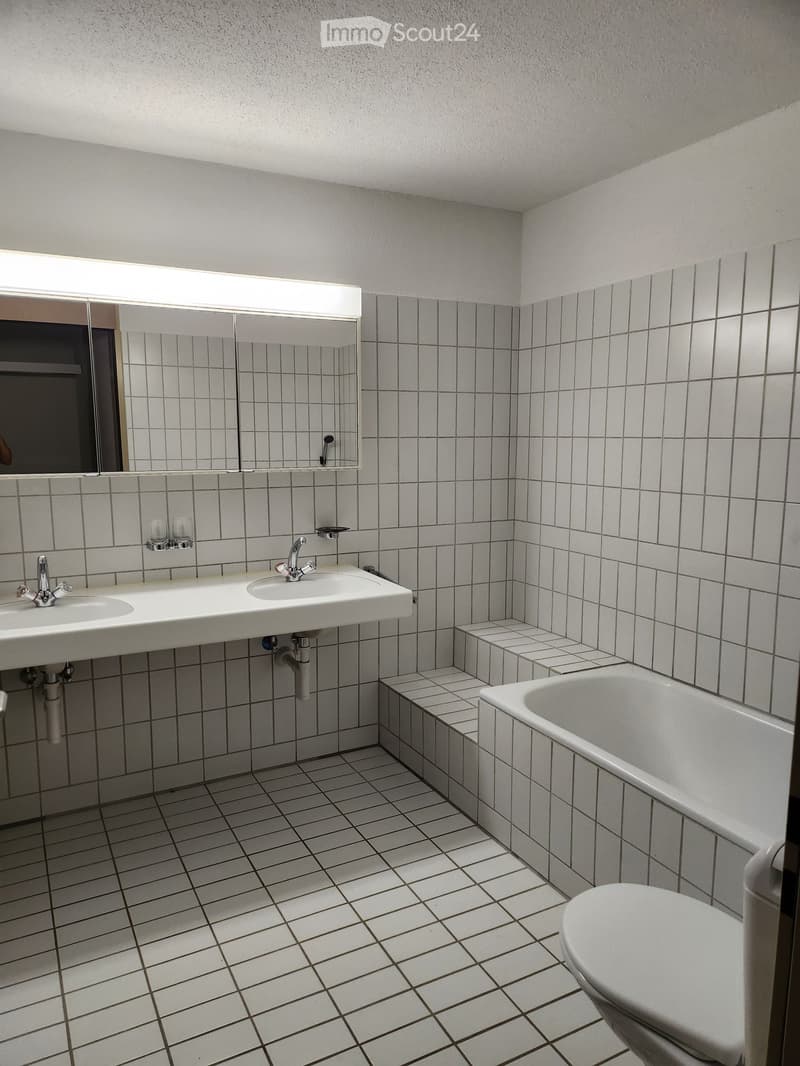 Bathroom with white tiled walls and floor, double sink, bathtub, toilet, and a large mirror above the sink.