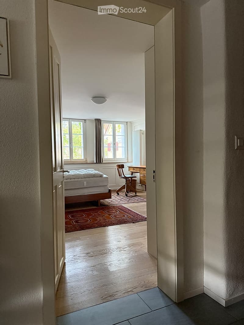 Bright bedroom with wooden flooring, large windows, a bed, a chair, a desk, and a patterned carpet.