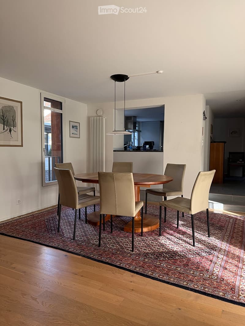 Dining room with a round wooden table surrounded by six beige chairs, a pendant light fixture, a radiator, and framed pictures on the wall. The room features wooden flooring and a patterned rug.