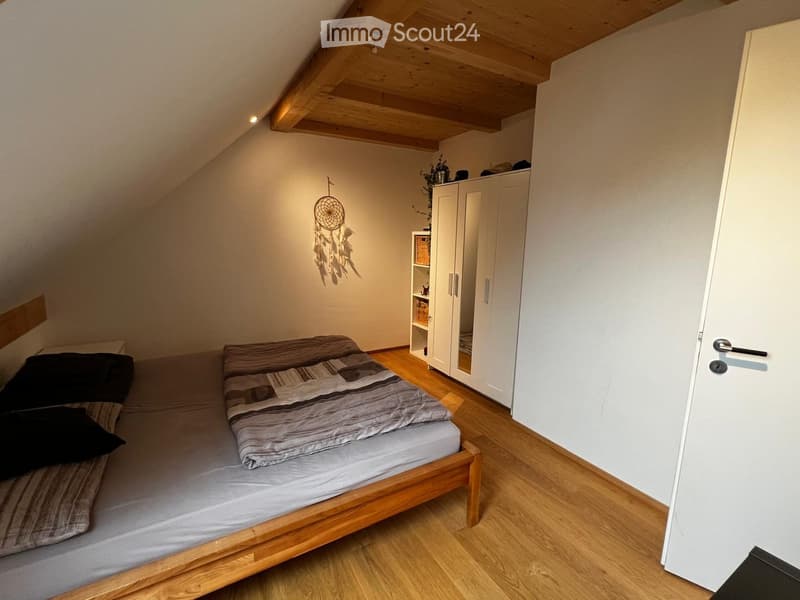 Cozy attic bedroom with wooden floors, slanted wooden ceiling, a single bed with grey bedding, a white wardrobe, a small shelf, and a dreamcatcher hanging on the wall.