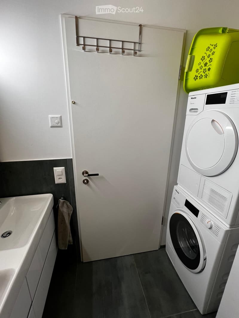 Bathroom with a bathtub, a white washing machine, a door, a towel hanging on a hook, and a small green storage bin on top of the washing machine.